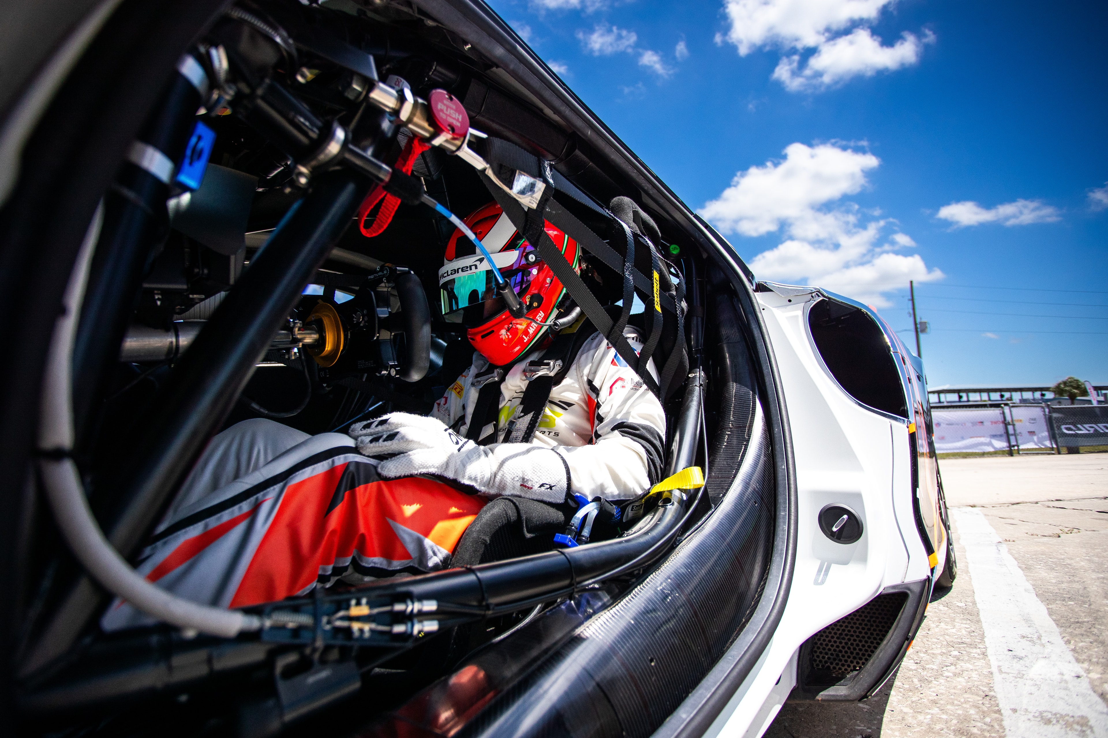 Driver strapped into the cockpit of a race car in pit lane, helmet on, waiting to head out.